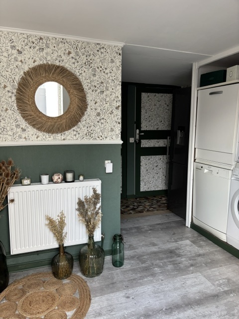L'entrée après les travaux : papier peint fleuri, soubassement vert foncé, miroir rond en fibre naturelle et sol stratifié clair.