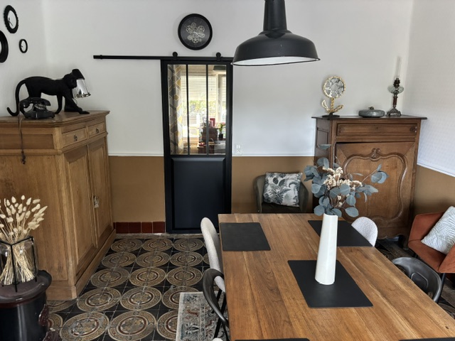 La salle à manger après les travaux : carreaux de ciment à motifs, verrière noire, grande table en bois et suspension d'atelier.